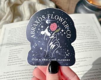 May include: A sticker with a dark blue background featuring the text "ABRAXOS FLOWER CO" and "STOP & SMELL THE FLOWERS". The sticker has a hand holding a pink rose, with stars and a crescent moon design.