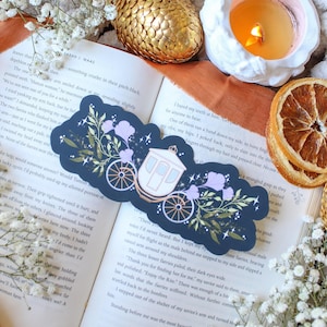 May include: A navy blue bookmark with a white carriage design, purple floral accents, and gold details. It rests on an open book with text, surrounded by white flowers and a golden decorative egg.