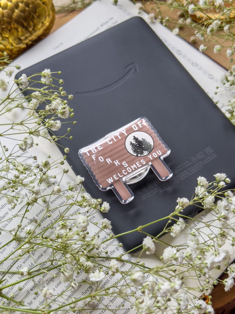 May include: A clear acrylic sign with the text "THE CITY OF FORKS WELCOMES YOU" sits on a black e-reader. The sign has a brown background with a tree graphic. White flowers and a book are in the background.