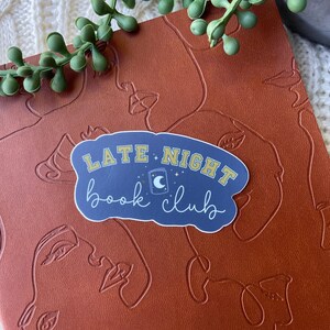 May include: A blue and yellow sticker with the text "Late Night Book Club" and a crescent moon and star design. The sticker is on a brown leather-like surface.