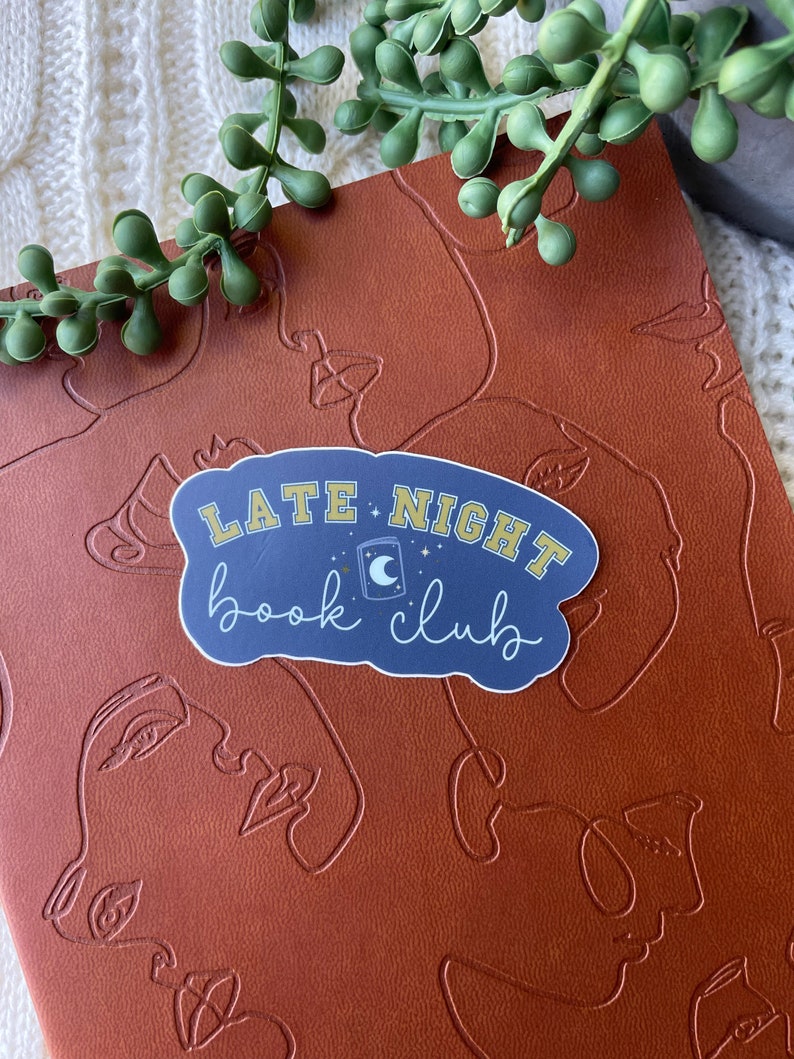 May include: A blue and yellow sticker with the text "Late Night Book Club" and a crescent moon and star design. The sticker is on a brown leather-like surface.