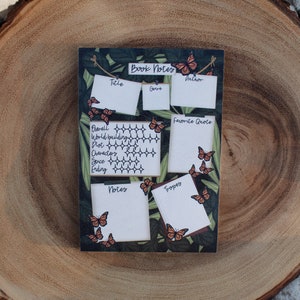 K&ouml;nnte beinhalten: Eine rechteckige Notiztafel f&uuml;r B&uuml;cher mit dunkelgr&uuml;nem Hintergrund und Schmetterlingsakzenten. Die Tafel hat Abschnitte f&uuml;r Titel, Genre, Autor, Lieblingszitat und Notizen. Die Tafel ist mit orangefarbenen Schmetterlingen verziert.