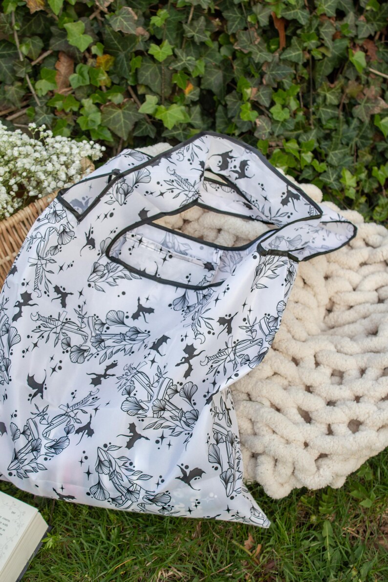 Peut inclure: Un sac de courses r&eacute;utilisable blanc avec un imprim&eacute; noir et blanc de chauves-souris, de fleurs et d'autres motifs d'inspiration gothique. Le sac est fait d'un mat&eacute;riau fin et translucide et a une bordure noire.