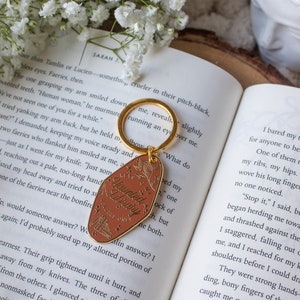 Puede incluir: Un llavero dorado con una etiqueta marr&oacute;n que dice "Keys to my Haunted Library, Readers Only". La etiqueta tiene un dise&ntilde;o floral y una peque&ntilde;a ilustraci&oacute;n de libros. El llavero est&aacute; sobre un libro abierto con flores blancas.