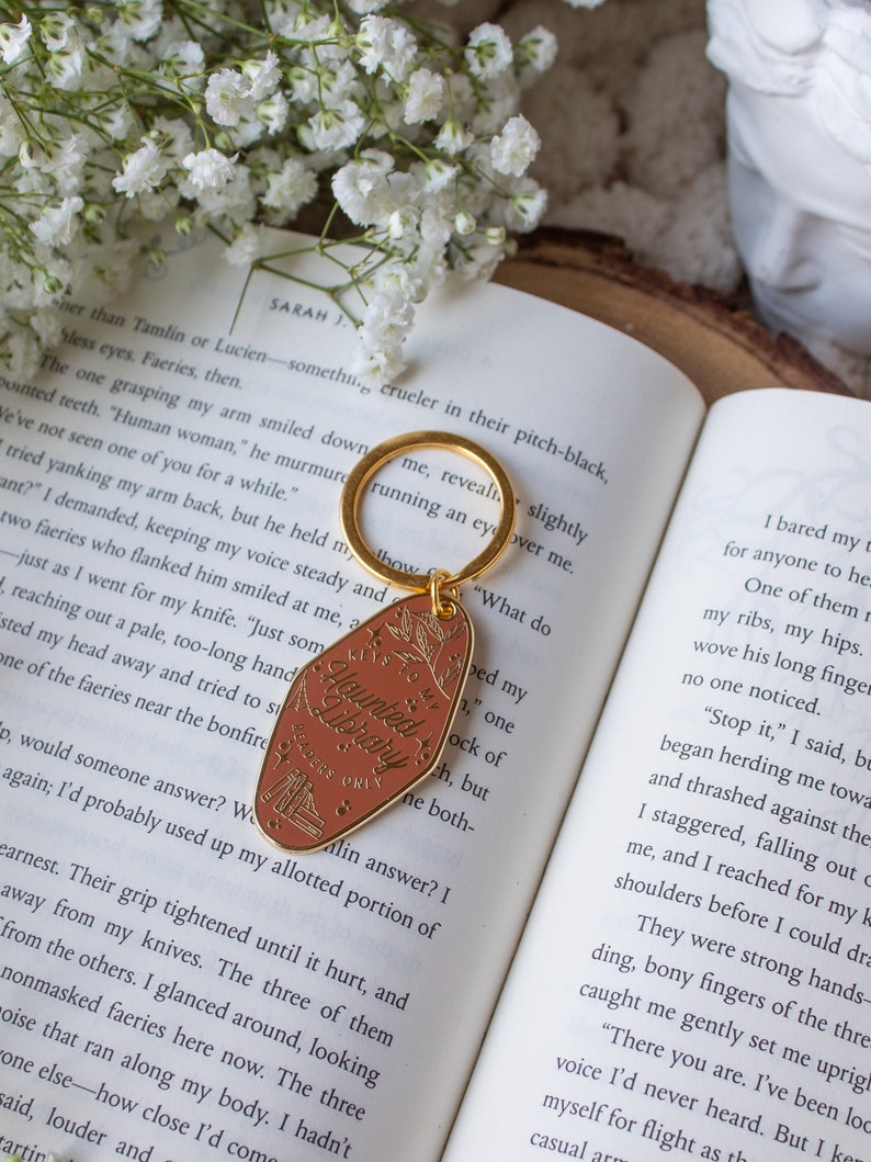 Puede incluir: Un llavero dorado con una etiqueta marr&oacute;n que dice "Keys to my Haunted Library, Readers Only". La etiqueta tiene un dise&ntilde;o floral y una peque&ntilde;a ilustraci&oacute;n de libros. El llavero est&aacute; sobre un libro abierto con flores blancas.