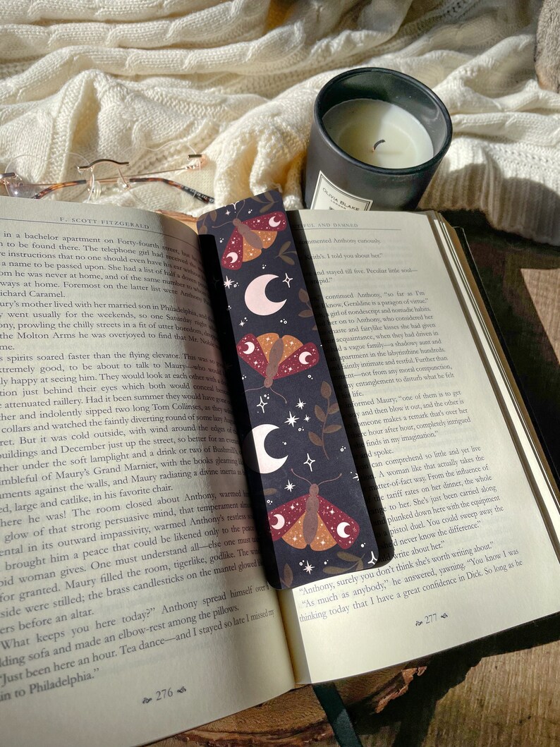 Puede incluir: Un marcap&aacute;ginas negro y marr&oacute;n con un dise&ntilde;o de luna y polilla. El marcap&aacute;ginas presenta una luna creciente con estrellas y una polilla con alas rojas, naranjas y marrones. El texto "Anthony, surely you don't think she's worth writing about?" est&aacute; impreso en el marcap&aacute;ginas.