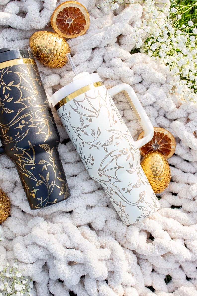 May include: Two stainless steel tumblers with a gold rim and a bat and vine design. The tumblers are on a white fuzzy blanket with gold decorative eggs and dried orange slices.