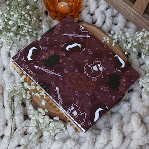Puede incluir: Una libreta de espiral color burdeos con un dise&ntilde;o blanco y dorado con calaveras, calderos y dagas. La libreta est&aacute; sobre una superficie blanca y esponjosa con un peque&ntilde;o portavelas naranja.