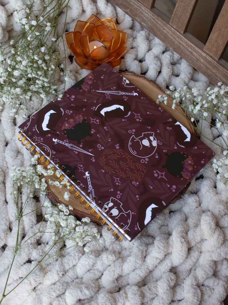 Puede incluir: Una libreta de espiral color burdeos con un dise&ntilde;o blanco y dorado con calaveras, calderos y dagas. La libreta est&aacute; sobre una superficie blanca y esponjosa con un peque&ntilde;o portavelas naranja.