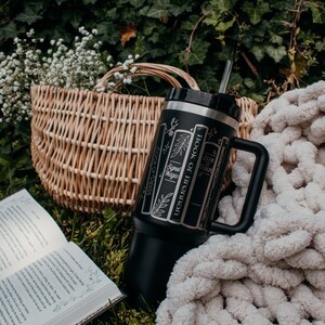 May include: A black stainless steel tumbler with a straw and a handle. The tumbler has a design of book spines with the titles "Book of Brennan", "Sigil Magic", "Book of Shadows", and "Book of Potions". The tumbler is sitting on a white blanket and green grass.
