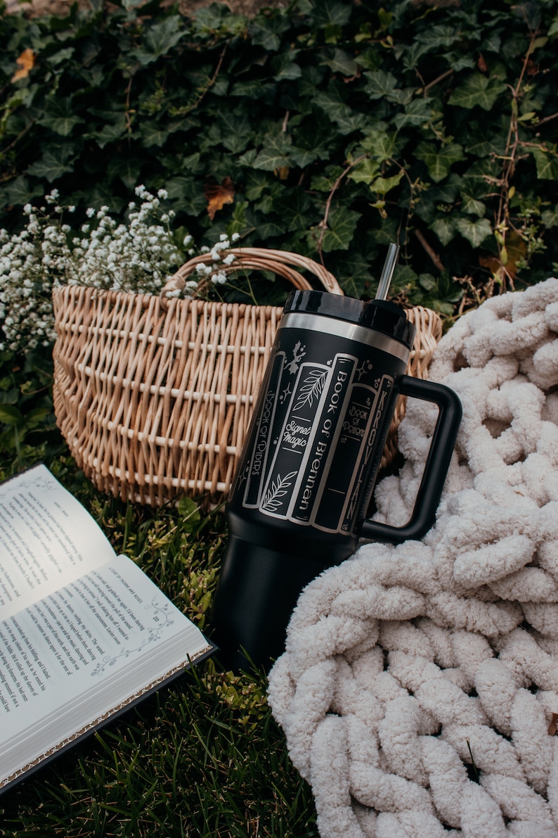 May include: A black stainless steel tumbler with a straw and a handle. The tumbler has a design of book spines with the titles "Book of Brennan", "Sigil Magic", "Book of Shadows", and "Book of Potions". The tumbler is sitting on a white blanket and green grass.