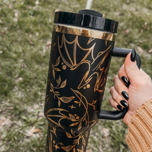 May include: Black and gold stainless steel tumbler with a bat and floral design. The tumbler has a black handle and a clear lid with a straw.