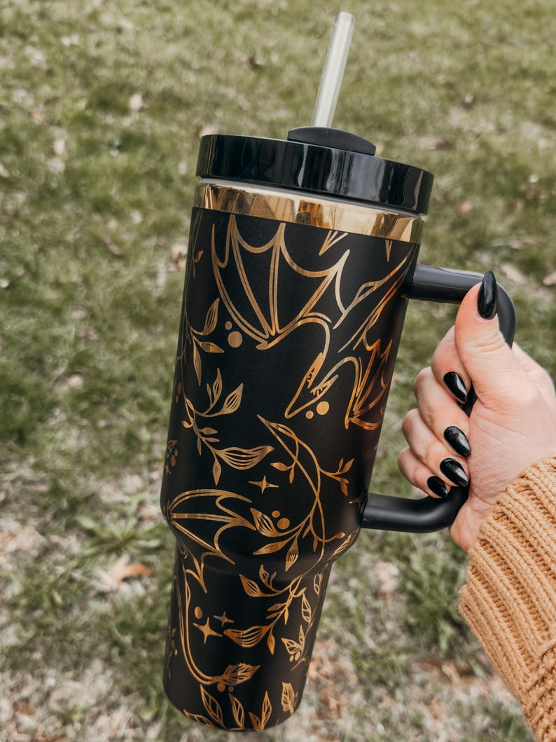 May include: Black and gold stainless steel tumbler with a bat and floral design. The tumbler has a black handle and a clear lid with a straw.