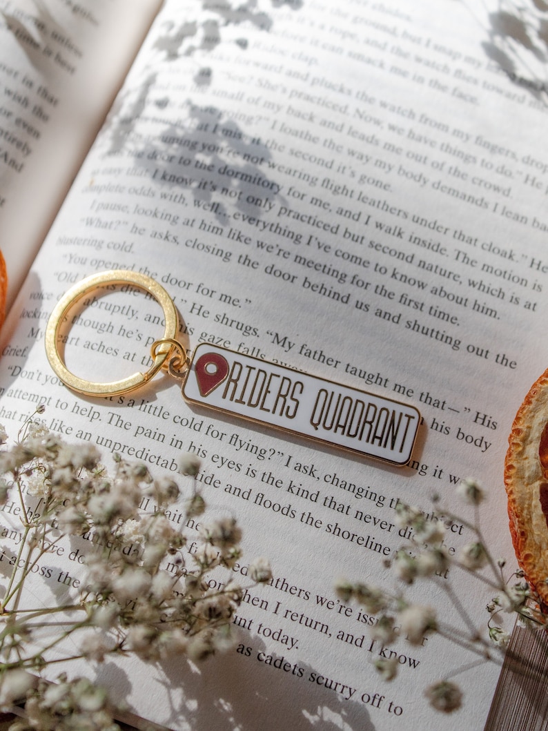 Op de afbeelding: Een gouden sleutelhanger met een rode en witte emaille hanger met de tekst "Riders Quadrant". De sleutelhanger ligt op een boek met witte bloemen en een schijfje sinaasappel.