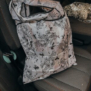 Peut inclure: Un sac de courses r&eacute;utilisable blanc avec un imprim&eacute; noir et blanc de diff&eacute;rents motifs, notamment des &eacute;toiles, des &eacute;p&eacute;es et des chauves-souris. Le sac est pli&eacute; et accroch&eacute; au dossier d'un si&egrave;ge de voiture.