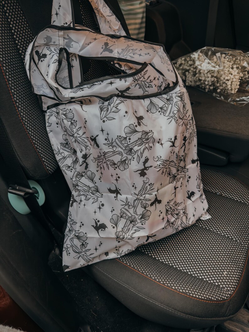 Peut inclure: Un sac de courses r&eacute;utilisable blanc avec un imprim&eacute; noir et blanc de diff&eacute;rents motifs, notamment des &eacute;toiles, des &eacute;p&eacute;es et des chauves-souris. Le sac est pli&eacute; et accroch&eacute; au dossier d'un si&egrave;ge de voiture.