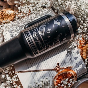 May include: A black stainless steel travel mug with a silver design featuring a dragon, a lemon, and stars. The mug has the text "Dragonkind" and "Riders Codex" on it. The mug is sitting on a book with a gold bookmark.