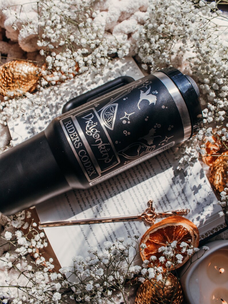 May include: A black stainless steel travel mug with a silver design featuring a dragon, a lemon, and stars. The mug has the text "Dragonkind" and "Riders Codex" on it. The mug is sitting on a book with a gold bookmark.