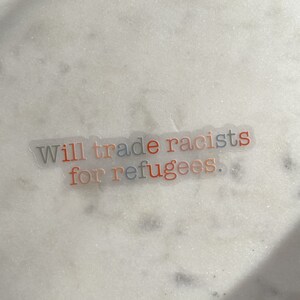 May include: A clear sticker with the text "Will trade racists for refugees." The words "racists" and "refugees" are in orange and blue, respectively. The sticker is on a white, speckled surface.