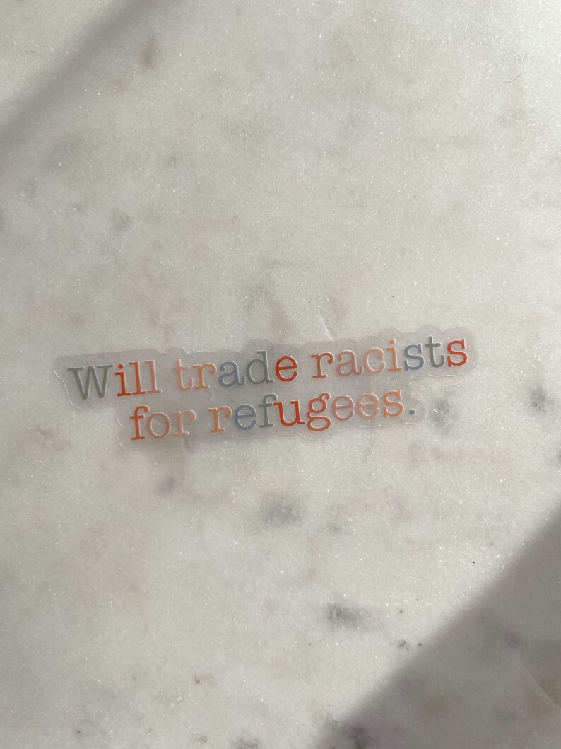 May include: A clear sticker with the text "Will trade racists for refugees." The words "racists" and "refugees" are in orange and blue, respectively. The sticker is on a white, speckled surface.