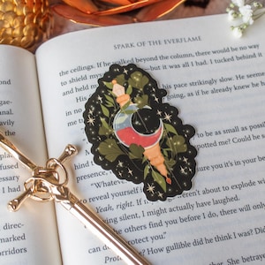 Puede incluir: Una pegatina con una botella de poción con un diseño de media luna, rodeada de hojas y estrellas, sobre un fondo negro. Un objeto decorativo dorado y un libro están en el fondo.