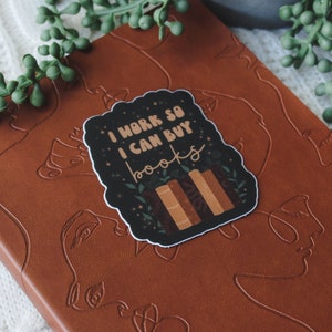 May include: A black and white sticker with the text "I work so I can buy books" and a stack of books with green leaves. The sticker is on a brown leather notebook with a line drawing of a face.
