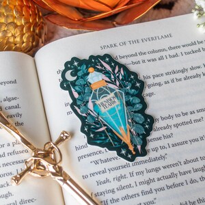 Puede incluir: Una pegatina con una botella de poción turquesa con el texto "HENRE'S TEARS" rodeada de hojas y una luna creciente. La pegatina está sobre un libro abierto con el título "SPARK OF THE EVERFLAME".