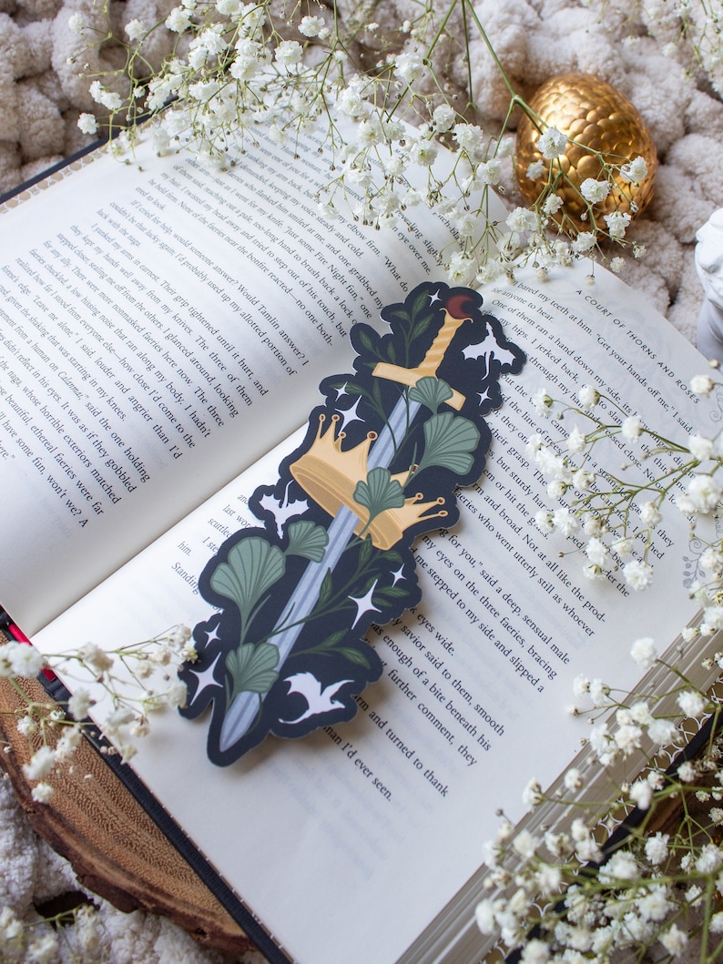 May include: A dark blue bookmark with a silver sword, gold crowns, and white birds. The sword is surrounded by green leaves and flowers. The bookmark is a great gift for book lovers.