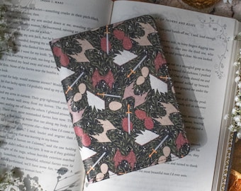 May include: A book sleeve featuring a dark pattern with repeating fantasy illustrations in white, red, and tan. The sleeve lies open on a book with visible text. The scene is decorated with additional elements.