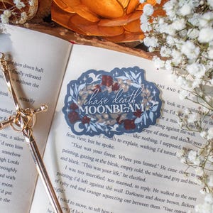 May include: A sticker with the text "chase death moonbeam" in a decorative font, surrounded by floral and leaf elements. The sticker is on an open book with a gold bookmark and a candle holder.