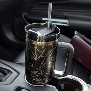 May include: A black and gold travel mug with a floral and bat design. The mug has a lid with a straw hole and a handle. The mug is in a car cup holder.