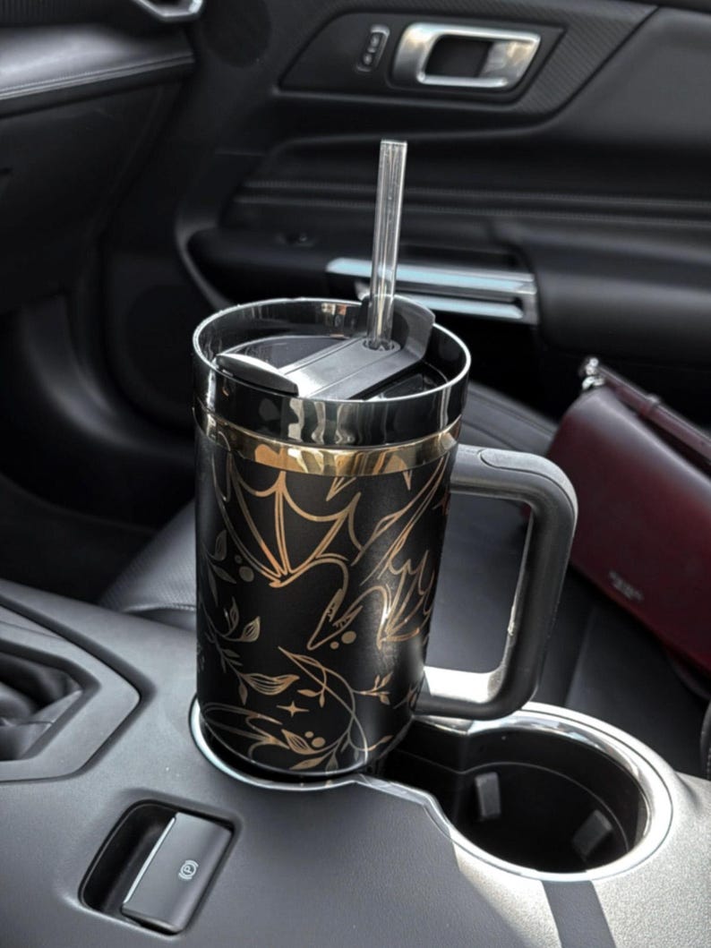 May include: A black and gold travel mug with a floral and bat design. The mug has a lid with a straw hole and a handle. The mug is in a car cup holder.