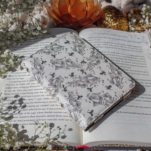 May include: A white and black patterned book cover featuring a floral and bird design, resting on an open book. The cover is rectangular and appears to be made of a soft material. The book is surrounded by white flowers and a decorative gold lotus.