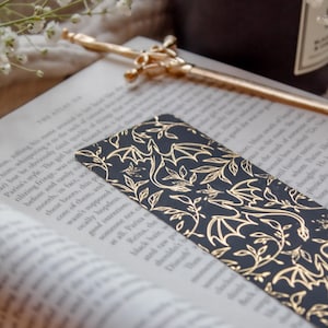 May include: A black bookmark with a gold leaf and dragon design rests on an open book. A gold pen with a sword-like design and a lit candle are also visible. The bookmark is a decorative accessory.