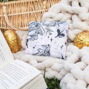 Peut inclure: Un sac fourre-tout en tissu blanc avec un motif floral noir et blanc. Le sac est pos&eacute; sur une couverture en tricot blanc avec un panier en osier et deux &oelig;ufs d&eacute;coratifs dor&eacute;s.