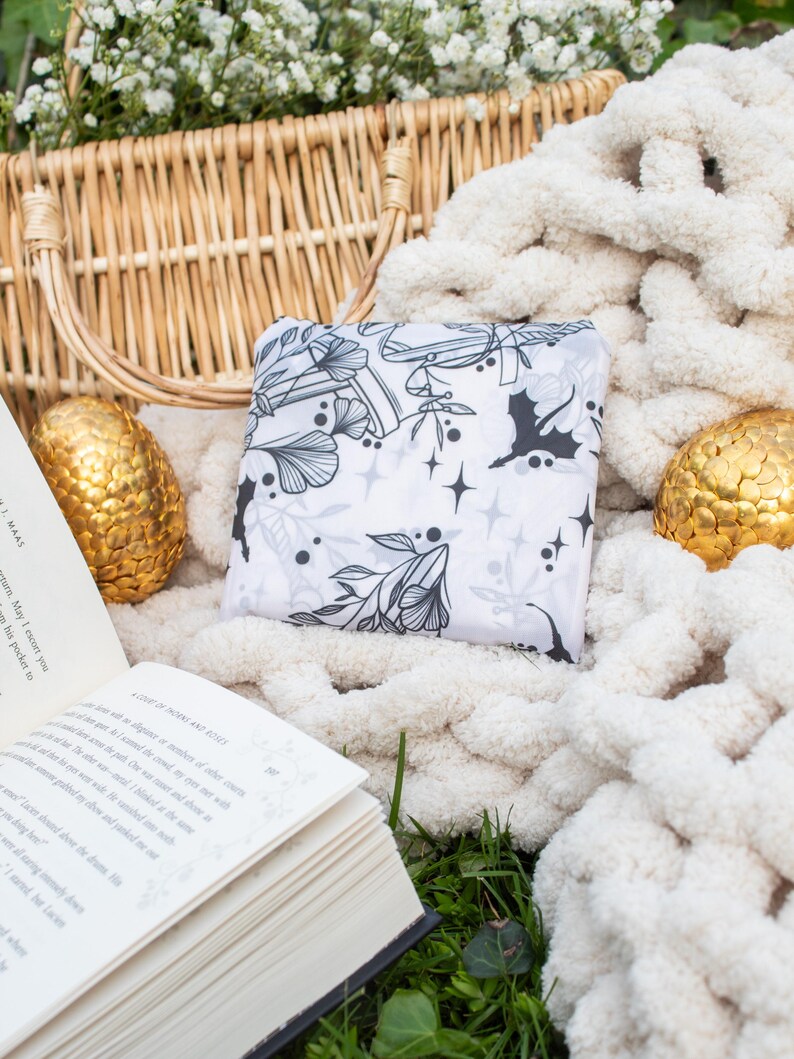 Peut inclure: Un sac fourre-tout en tissu blanc avec un motif floral noir et blanc. Le sac est pos&eacute; sur une couverture en tricot blanc avec un panier en osier et deux &oelig;ufs d&eacute;coratifs dor&eacute;s.