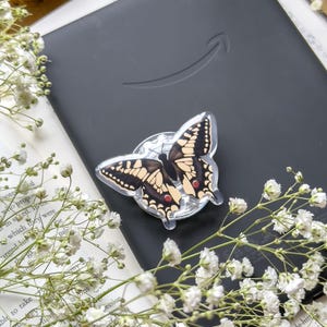 May include: A clear butterfly-shaped phone grip with black and tan wings and red spots is attached to a black e-reader. White flowers and a book are in the background. The e-reader has the Amazon logo.