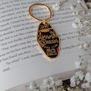 Puede incluir: Llavero negro y dorado del Club de Lectura Horror Reader. El llavero presenta un fondo negro con detalles dorados, un gráfico de cuchillo y el texto "Horror Reader Book Club."