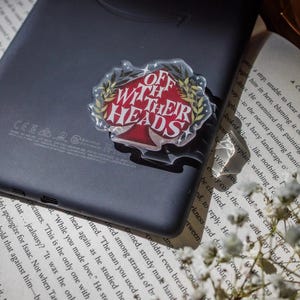May include: A clear acrylic pin with the phrase "Off With Their Heads" in red and white, surrounded by a gold laurel wreath. The pin is on a black electronic device.