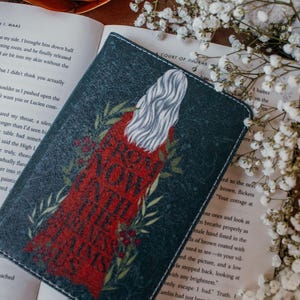 Puede incluir: Una cubierta de libro verde oscuro con un diseño que presenta una figura con un vestido rojo y cabello blanco, con las palabras "FROM NOW UNTIL THE END" en rojo. La cubierta está sobre un libro abierto con flores blancas.