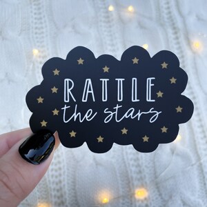 Peut inclure: Autocollant noir en forme de nuage avec des &eacute;toiles dor&eacute;es et le texte "RATTLE the stars" en blanc.