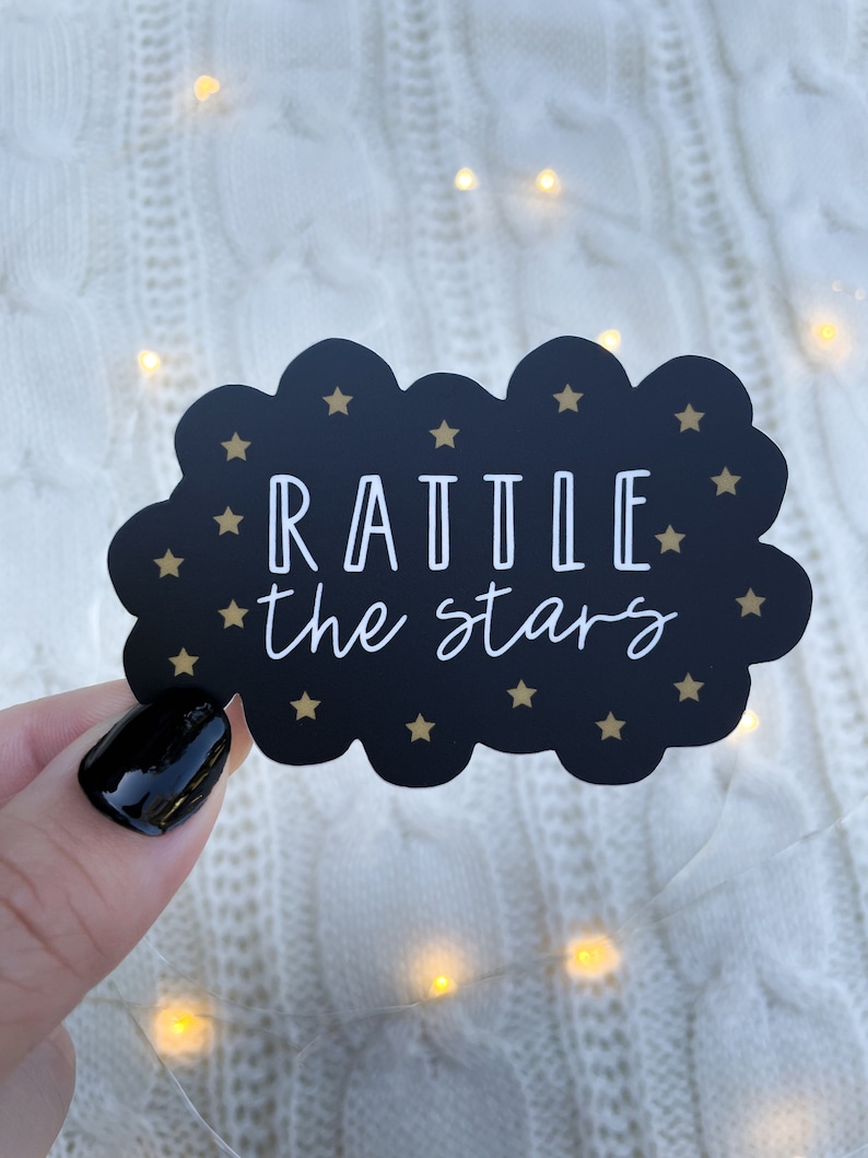 Peut inclure: Autocollant noir en forme de nuage avec des &eacute;toiles dor&eacute;es et le texte "RATTLE the stars" en blanc.