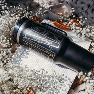 May include: A black stainless steel tumbler with a white design featuring the text "Book of Riders" and "Riders Quadrant Rulebook". The tumbler is sitting on a book with white flowers and dried orange slices.