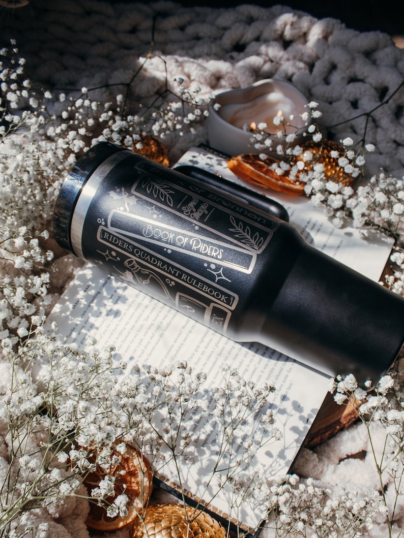 May include: A black stainless steel tumbler with a white design featuring the text "Book of Riders" and "Riders Quadrant Rulebook". The tumbler is sitting on a book with white flowers and dried orange slices.