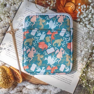 May include: Teal, zippered tablet sleeve with a repeating pattern of dragons, books, and foliage in shades of peach, gold, and white. The sleeve is rectangular with a white border and a front pocket.