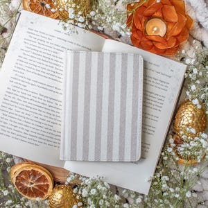 May include: A book with open pages, a striped gray and white notebook, and decorative elements like dried orange slices and golden eggs. A candle burns in an orange flower-shaped holder, creating a cozy atmosphere.