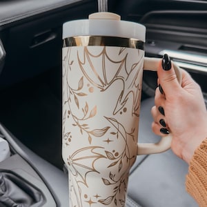 May include: A white and gold travel mug with a floral and bat design. The mug has a lid and a straw. It is sitting in a car cup holder.