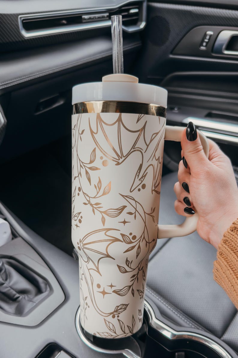 May include: A white and gold travel mug with a floral and bat design. The mug has a lid and a straw. It is sitting in a car cup holder.