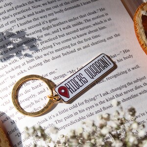 Op de afbeelding: Een gouden sleutelhanger met een rode en witte emaille pin die "Riders Quadrant" zegt. De pin heeft een rood pin-icoon. De sleutelhanger ligt op een boek met witte bloemen en een stuk oranje citrusvrucht.