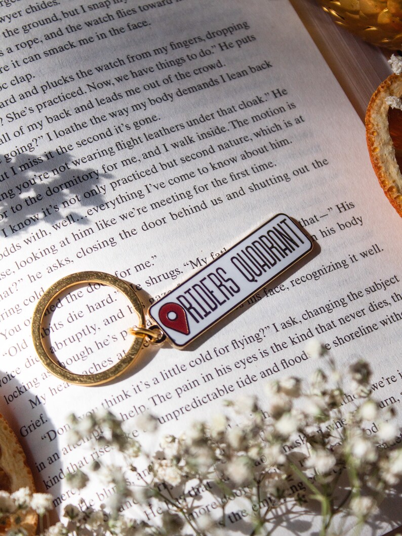 Op de afbeelding: Een gouden sleutelhanger met een rode en witte emaille pin die "Riders Quadrant" zegt. De pin heeft een rood pin-icoon. De sleutelhanger ligt op een boek met witte bloemen en een stuk oranje citrusvrucht.